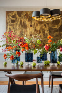 Flowers in vases and apples on a wooden table under a modern chandelier with gold art behind it